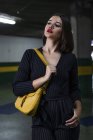Femme à la mode en robe noire avec rouge à lèvres rouge et petit sac jaune debout sur un parking — Photo de stock