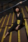 Femme à la mode en robe noire avec rouge à lèvres rouge et petit sac jaune assis à la main courante de l'escalier dans une rue de la ville au crépuscule — Photo de stock