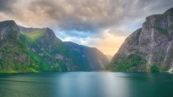 Потрясающий пейзаж на зеленой воде, отражающей облачное небо, омывающий скалистые горы зеленым деревом и травой в Норвегии — стоковое фото