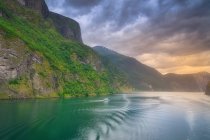 Boat in stunning landscape on green water reflecting cloudy sky washing rocky mountains with green tree and grass in Norway — Stock Photo