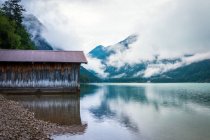 Remise pour bateaux située près de l'eau tranquille d'un étang près d'une crête montagneuse par une journée nuageuse en Autriche — Photo de stock