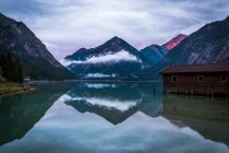 Shabby shed for boats located near tranquil water of pond near mountain ridge on cloudy day in Austria — Stock Photo