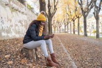 African American woman in yellow hat and warm jacket using smartphone sitting on road with autumn leaves in park — Stock Photo