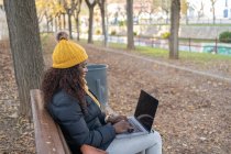 Vista lateral da moda alegre mulher afro-americana em chapéu amarelo e casaco quente trabalhando no laptop confortavelmente sentado no banco de madeira no parque de outono — Fotografia de Stock