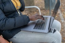 Афроамериканська жінка в жовтому капелюсі і теплий піджак працюють на ноутбуці зручно сидячи на дерев'яній лавці восени парку — стокове фото