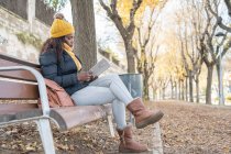 Vista lateral da mulher afro-americana elegante pensativo em chapéu amarelo e casaco quente confortável sentado no banco de madeira e livro de leitura no parque de outono — Fotografia de Stock