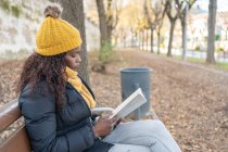 Vista lateral da mulher afro-americana elegante pensativo em chapéu amarelo e casaco quente confortável sentado no banco de madeira e livro de leitura no parque de outono — Fotografia de Stock