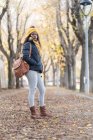 Entusiasta donna afroamericana alla moda in cappello giallo e giacca calda con zaino in pelle e mani in tasca guardando la fotocamera con sorriso sul parco stradale autunnale — Foto stock