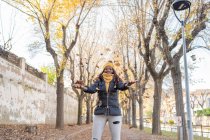 Стильная, восторженная афроамериканка в теплой куртке весело бросает сухие осенние листья в воздух в парке — стоковое фото