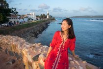 Satisfied Asian woman on vacation in colorful light dress sitting on rocked fence of tropical quay and looking away at Galle at Sri Lanka — Stock Photo