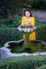 Щаслива жінка з Азії посміхається й торкається чистої води фонтану, коли відпочиває в саду в Малазі (Іспанія). — стокове фото