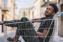 Вид сбоку на застенчивого юношу-афроамериканца в повседневной одежде, смотрящего в камеру, сидя в металлическом торговом тролли на улице — стоковое фото