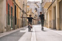Un jeune Afro-américain se promène dans un scooter électrique alors qu'un homme noir conduit une bicyclette dans la rue en regardant une caméra — Photo de stock
