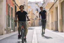 Alegre jovem africano americano homem equitação scooter elétrico enquanto preto masculino está dirigindo bicicleta na rua olhando para câmera — Fotografia de Stock