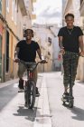 Веселый молодой афроамериканец ездит на электрическом скутере, в то время как чернокожий мужчина ездит на велосипеде по улице, глядя в камеру — стоковое фото