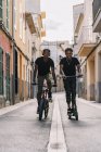 Un joven y alegre hombre afroamericano que monta un scooter eléctrico mientras el hombre negro conduce en bicicleta por la calle mirando la cámara. - foto de stock