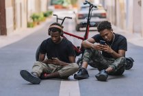 Веселий молодий афроамериканець, який ділиться фотографіями на мобільних телефонах з приємним чорним хлопцем у навушниках. — стокове фото