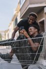 Glücklicher junger afroamerikanischer Mann in lässiger Kleidung sitzt im Einkaufswagen und teilt Nachrichten auf dem Smartphone mit einem schwarzen männlichen Freund in Kopfhörern beim gemeinsamen Zeitvertreib auf der Straße — Stockfoto
