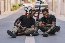 Щасливий молодий афроамериканець, який ділиться фотографіями на мобільному телефоні з радісним чорним другом у навушниках. — стокове фото