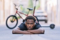 Concentrated youthful African American man wearing headphones listening to music while lying on asphalt road in street — Stock Photo