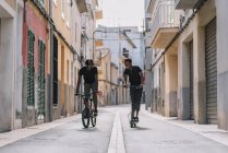 Allegro giovane afroamericano uomo in sella scooter elettrico mentre nero maschio amico è in bicicletta di guida in strada — Foto stock