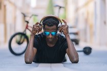 Concentrated youthful African American man in sunglasses wearing headphones and listening to music while lying on asphalt road in street — Stock Photo