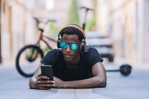 Concentrated youthful African American man in sunglasses wearing headphones and listening to music on the mobile phone while lying on asphalt road in street — Stock Photo