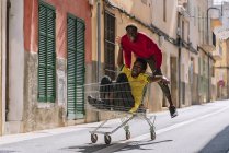 Happy carefree young African American friends in casual clothes riding around in shopping trolley in street — Stock Photo