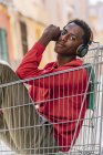 Seitenansicht eines nachdenklichen, jugendlichen afroamerikanischen Teenagers in lässiger Kleidung, der in einem Einkaufswagen aus Metall auf der Straße in die Kamera blickt — Stockfoto