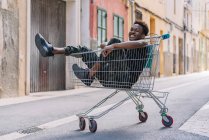 Vista lateral del joven y penoso adolescente afroamericano con ropa casual mirando a la cámara mientras estaba sentado en un carro de compras de metal en la calle. - foto de stock