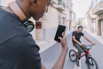 Desde arriba, el joven afroamericano enfocado en el cultivo tomó fotos en teléfonos inteligentes de un amigo negro masculino en bicicleta en la calle. - foto de stock