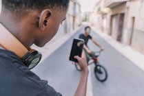 From above crop focused young African American guy taking picture on smartphone of male black friend on bike in street — Photo de stock