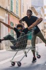 Fröhliche unbeschwerte junge afroamerikanische Freunde in lässiger Kleidung, die im Einkaufswagen auf der Straße herumfahren — Stockfoto