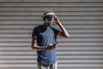 Elegante homem americano Africano moderno em óculos de sol e fones de ouvido ouvir música no telefone celular na parede listrada na rua — Fotografia de Stock
