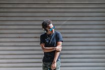 Elegante homem americano Africano moderno em óculos de sol e fones de ouvido ouvir música na parede listrada na rua — Fotografia de Stock