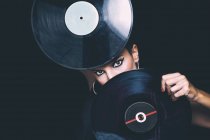 Selbstbewusste junge Frau mit hellem Eyeliner, die in die Kamera blickt und ihr Gesicht hinter Schallplatten im Studio auf schwarzem Hintergrund versteckt — Stockfoto