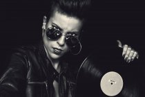 Angry confident female rocker in leather jacket and aviator sunglasses looking at camera and holding vinyl record in studio on black background — Stock Photo