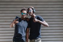Cool jóvenes adolescentes afroamericanos en gafas de sol tomando fotos con teléfono móvil mientras se encuentran bajo la luz del sol en la calle. - foto de stock