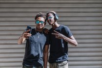 Cool youthful African American male teenagers in sunglasses taking picture with mobile phone while standing in sunlight in street — Stock Photo