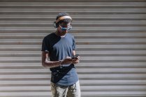 Stilvoller, moderner Afroamerikaner mit Sonnenbrille und Kopfhörer, der an der gestreiften Wand in der Straße Musik auf dem Handy hört — Stockfoto