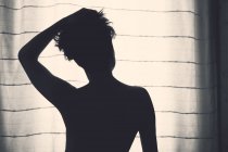 Visão traseira de silhueta irreconhecível de fêmea com cabelo curto levantando a mão para cima e de pé na frente da janela com cortina — Fotografia de Stock