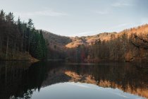 Paisaje escénico con bosque verde y amarillo y colinas reflejadas en el tranquilo agua oscura del hermoso lago en el día soleado - foto de stock