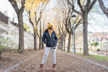 Moda encantadora afro-americana mulher em chapéu amarelo e casaco quente com as mãos no bolso na estrada com folhas de outono no parque — Fotografia de Stock