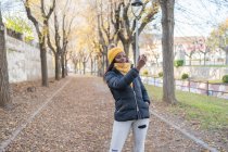Cheerful fashion African American woman in yellow hat and warm jacket taking selfie on smartphone on road with autumn leaves in park — Stock Photo