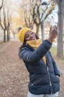 Cheerful fashion African American woman in yellow hat and warm jacket taking selfie on smartphone on road with autumn leaves in park — Stock Photo