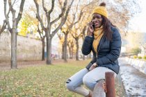 Cheerful relaxed African American woman in yellow hat and warm jacket speaking on smartphone sitting on wooden fence with autumn leaves in park — Stock Photo