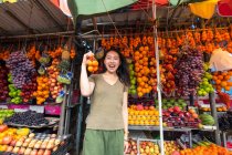 Emocionado turista asiático en ropa casual riendo mientras sostiene bolsillo con mandarinas en colorido mercado al aire libre en Sri Lanka - foto de stock