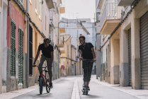 Веселий молодий афроамериканець їде на електричному скутері, а чорний чоловік їздить на велосипеді на вулиці. — стокове фото