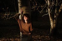 Pleased curly haired woman in casual wear touching autumn yellow leaf and smiling in woods — Stock Photo