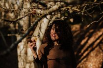 Приятно кудрявая женщина в повседневной одежде касаясь осенью желтый лист и улыбаясь в лесу — стоковое фото